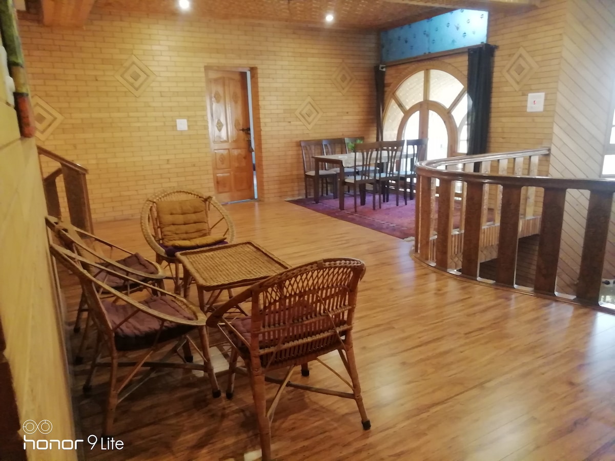 A spacious area features wooden flooring and a warm color palette. Seating arrangements include wicker chairs and a small table, while a dining table is visible in the background. Large windows allow natural light to enter, contributing to an open and airy feel.