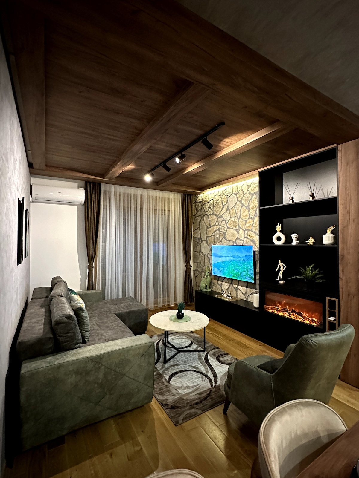 A cozy living area features a sectional sofa and a plush armchair, with a round coffee table at the center. A wall-mounted TV is complemented by an electric fireplace beneath it. Natural light filters through sheer curtains, illuminating the stone-accented wall.