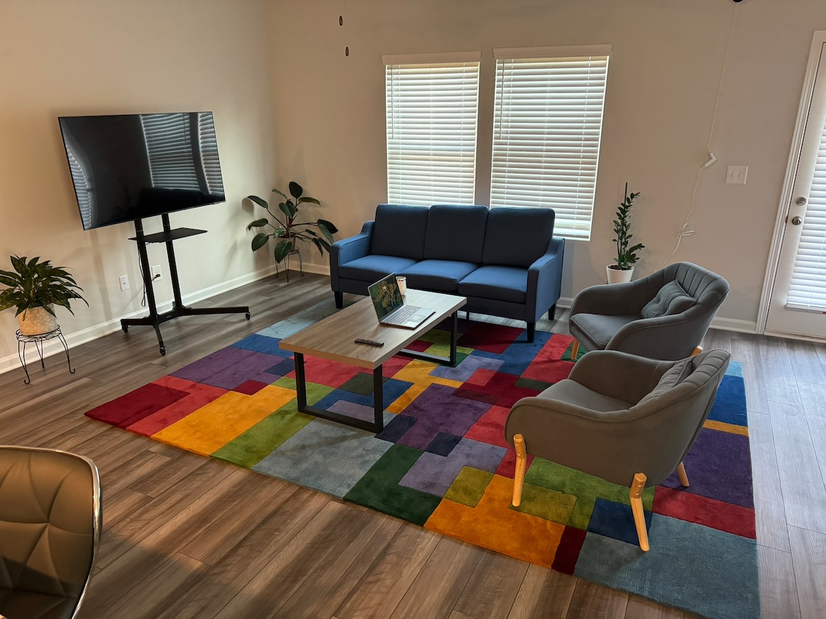 A comfortable living area features a modern blue sofa and two grey armchairs arranged around a central coffee table. A colorful area rug covers the floor, adding vibrancy to the space. Natural light enters through the multi-pane windows, creating an airy atmosphere.