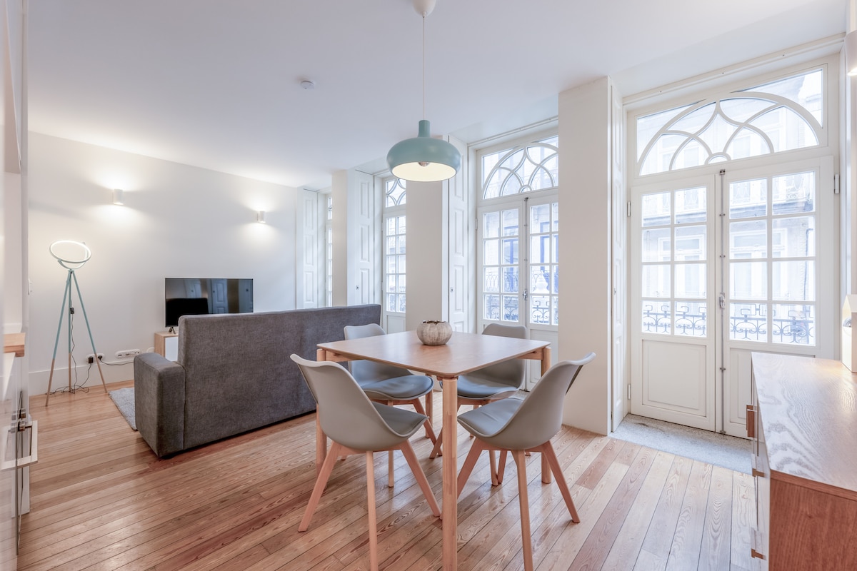 A spacious living area is illuminated by natural light flowing through large, arched windows. The room features a comfortable sofa, a wooden dining table surrounded by four chairs, and a modern floor lamp. Light wood flooring enhances the bright ambiance of the space.