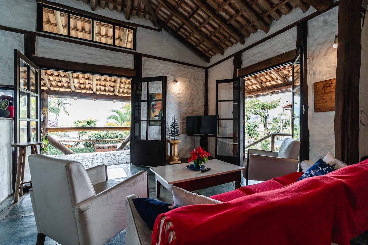 A rustic living area features a cozy seating arrangement with white armchairs and a wooden table. Natural light floods in from large glass doors leading to a balcony, showcasing views of the surrounding greenery. The charm of exposed wooden beams adds warmth to the ambiance.