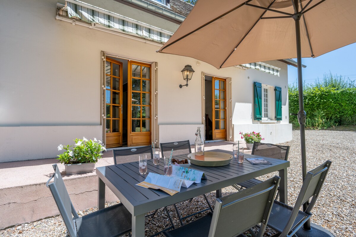 An outdoor dining area is positioned under a large umbrella, featuring a rectangular table surrounded by several chairs. Glasses of water and a decorative platter rest on the table. The backdrop includes French doors leading to the house, framed by green plants.