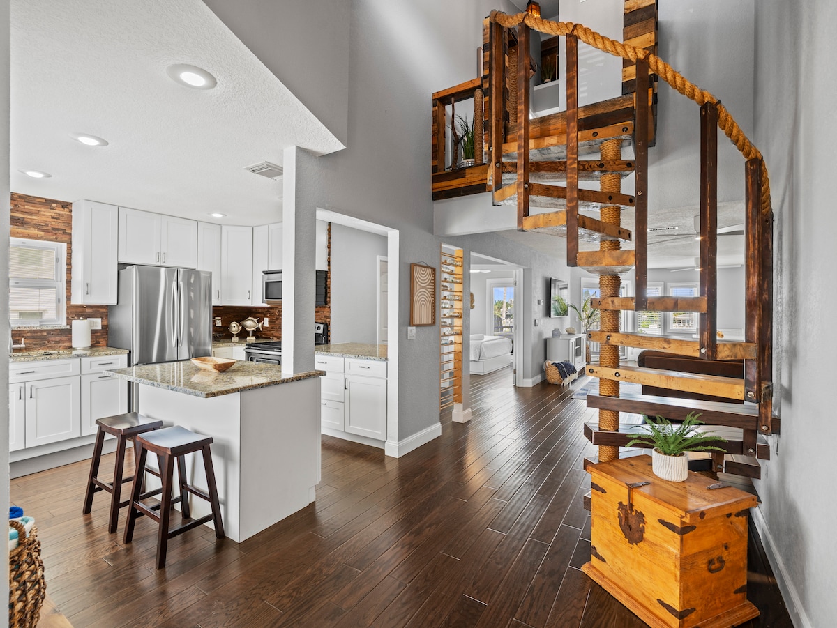 An open-concept living space is illuminated by natural light, featuring a modern kitchen with stainless steel appliances and a granite island. A spiral staircase is prominently displayed, leading to the upper floor, while wooden accents and decorative elements contribute to the welcoming environment.