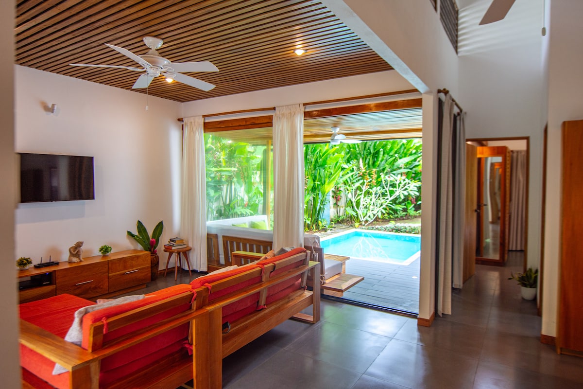 The living area features light wood accents and a bright red sofa, providing a welcoming space. Large sliding doors open to the pool area, surrounded by lush greenery. A wall-mounted television is visible, and ceiling fans contribute to the airy atmosphere.