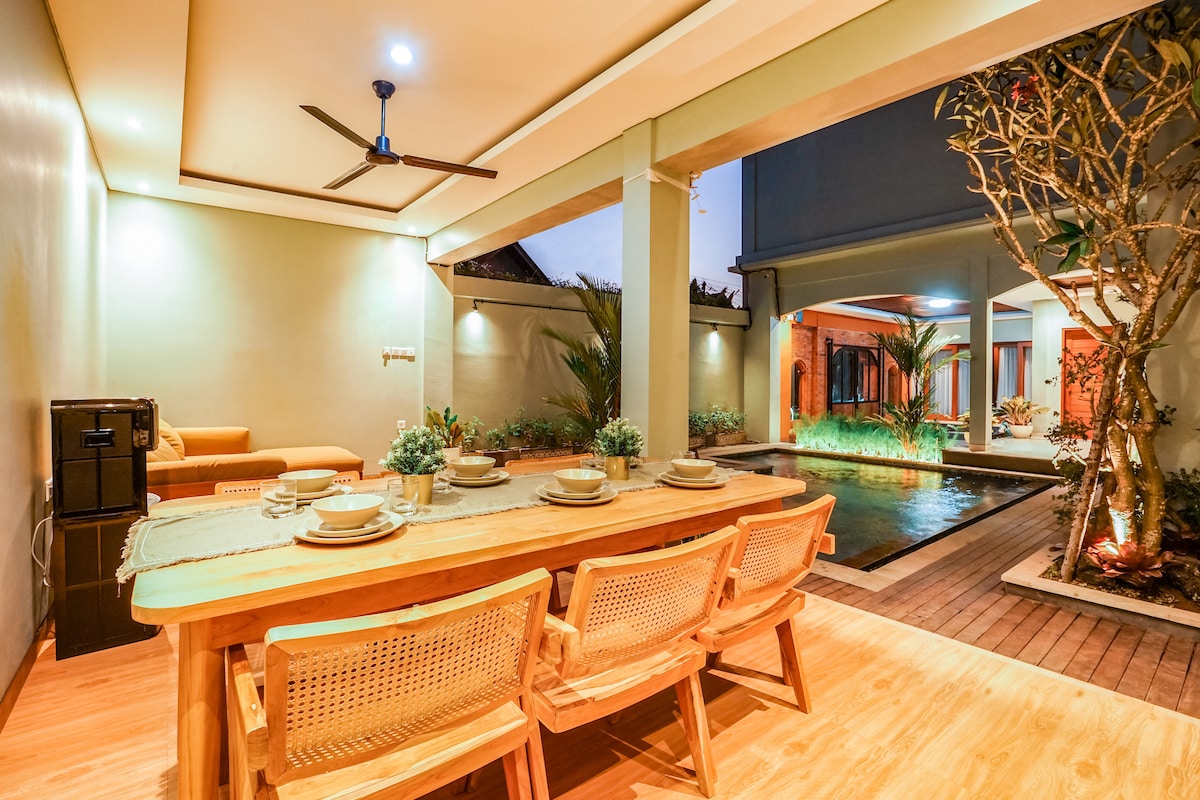 A spacious communal area features a wooden dining table with seating for six, complemented by a refreshing view of the adjacent swimming pool. Potted plants and a serene water feature enhance the tranquil atmosphere, while a ceiling fan offers a cooling breeze.