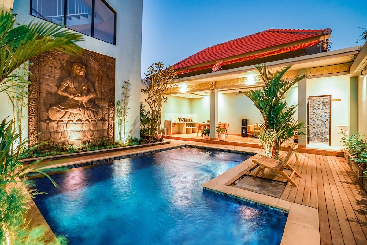 A serene outdoor space features a tranquil swimming pool surrounded by lush greenery and wooden decking. A detailed wall sculpture of a Buddha stands nearby, accentuating the calming atmosphere. The communal area showcases comfortable seating, enhancing the inviting environment for relaxation and socializing.