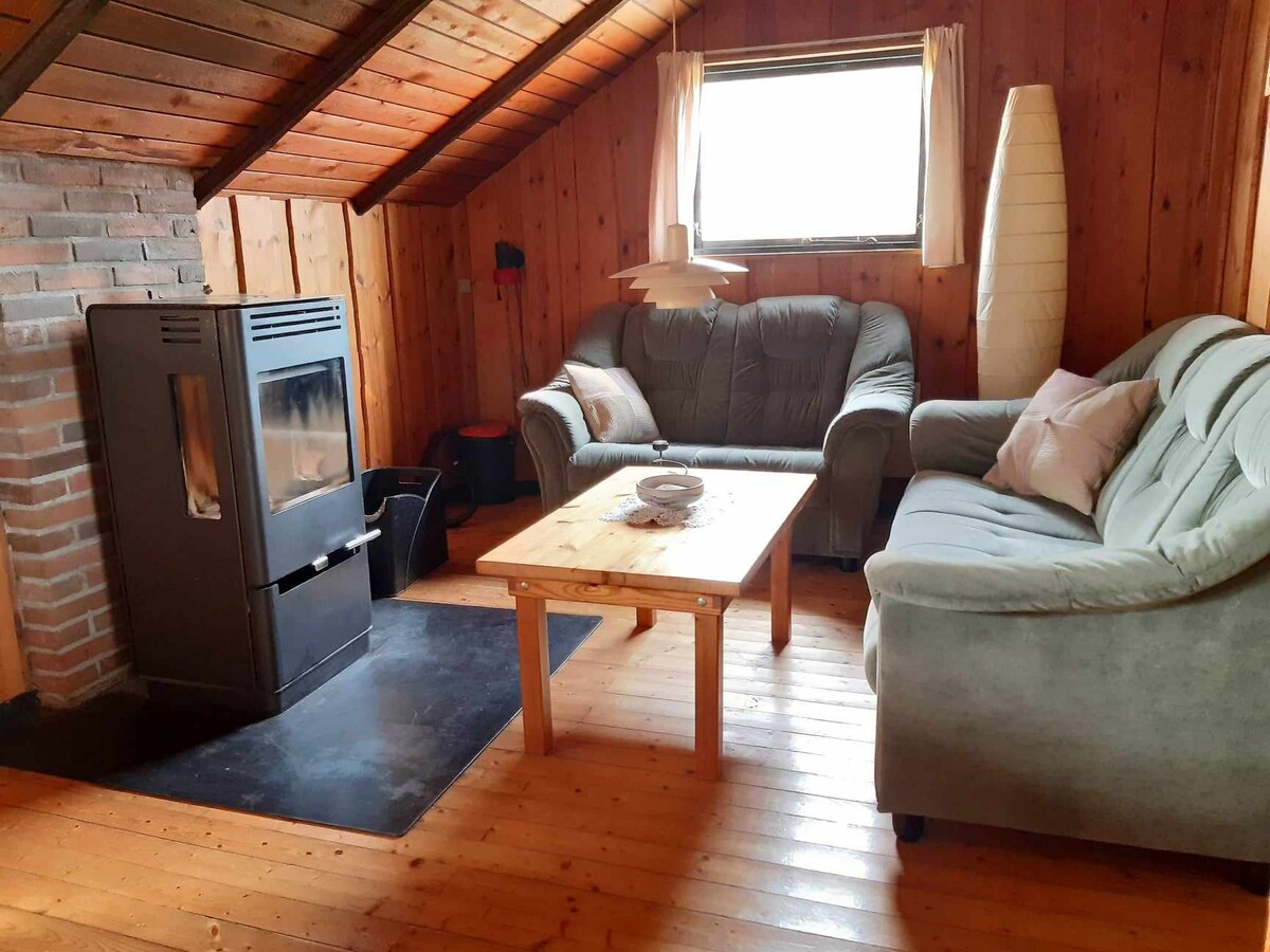 A cozy seating area is present, featuring a light blue sofa and a gray couch, positioned around a wooden coffee table. A warm fireplace with a black casing stands against a brick wall, with natural light flowing in from a window adorned with sheer curtains.