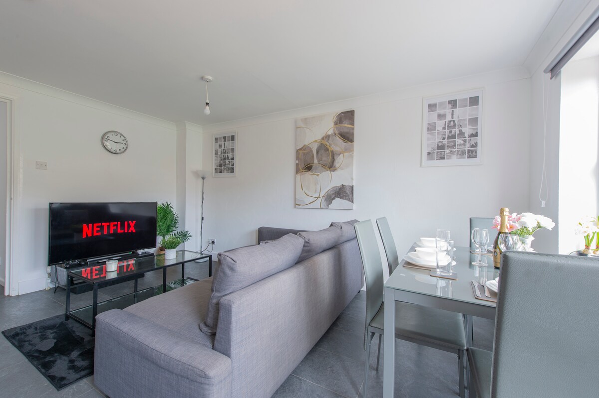 A modern living area is presented with a comfortable grey sofa facing a black flat-screen television displaying the Netflix logo. A glass dining table with seating for four is visible, adorned with dining ware and a small vase of flowers. Artwork complements the walls.