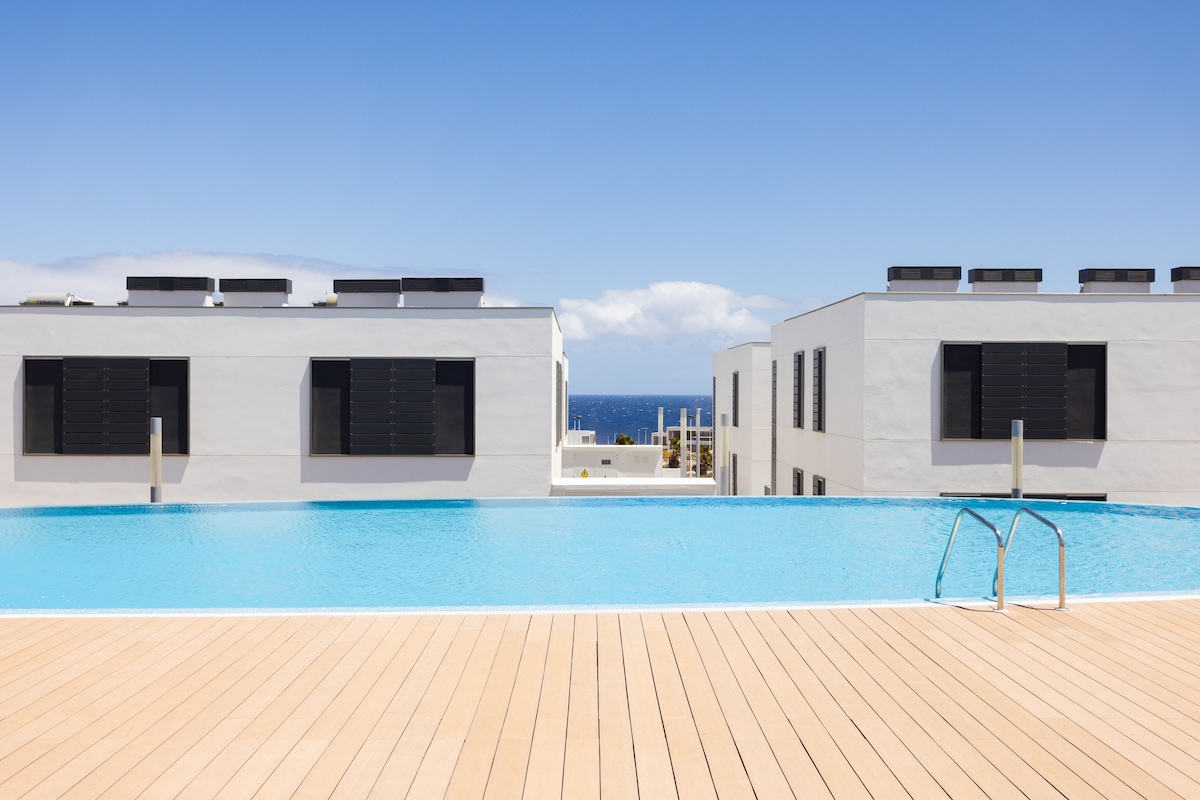 A serene infinity pool is displayed against a backdrop of modern white buildings. The pool's calm blue water reflects the clear sky, while wooden decking provides a warm contrast. The horizon reveals the ocean, offering a picturesque view of the coastal surroundings.