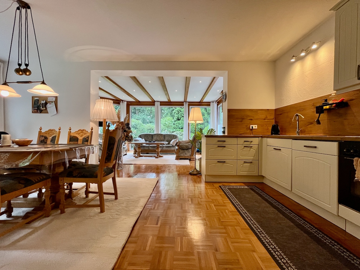 An open-plan kitchen area is visible, featuring light cabinetry and modern appliances. A dining table with six chairs is centrally positioned, illuminated by soft lighting. Through the archway, a living area can be seen, highlighted by natural light from large windows overlooking greenery.