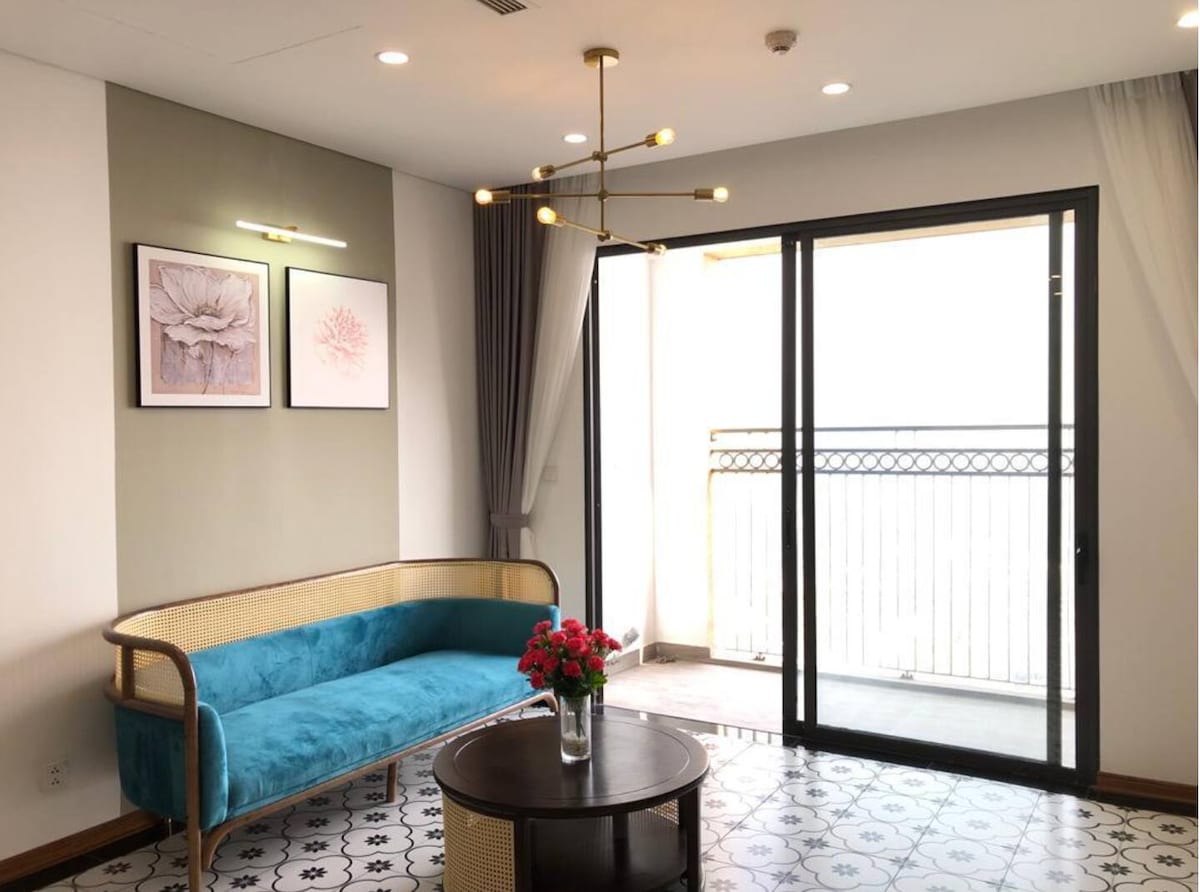 A bright living area showcases a blue upholstered sofa paired with a round dark table, accented by fresh flowers. Two floral art pieces adorn the wall, and sliding glass doors open to a balcony, allowing natural light to flow in.