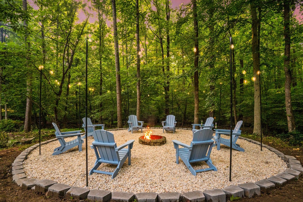 A circle of blue Adirondack chairs surrounds a fire pit, set on a gravel surface amidst a lush forest. Soft lighting from string lights overhead enhances the serene outdoor setting, inviting relaxation and conversation around the fire.