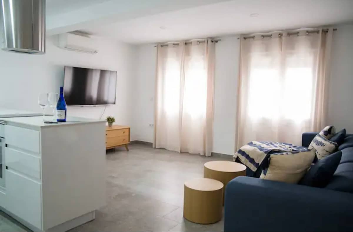 A bright living area features a comfortable sofa adorned with decorative pillows. A stylish wooden coffee table complements the space, while a television is mounted on the wall. Natural light enters through sheer curtains that frame the windows.