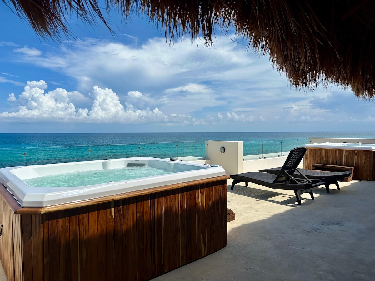 A sunlit outdoor space features a wooden hot tub and a comfortable lounge chair. The area is adorned with a thatched roof, providing shade. The expansive ocean view reveals clear turquoise waters and a blue sky adorned with soft clouds.