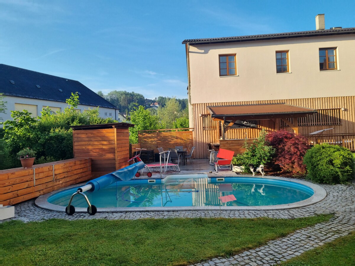 A tranquil outdoor space features a sparkling pool surrounded by lush greenery and a stone path. Deck chairs and an outdoor dining area are visible under a shaded structure. The nearby building showcases a wooden fence and soft sunlight illuminates the scene.