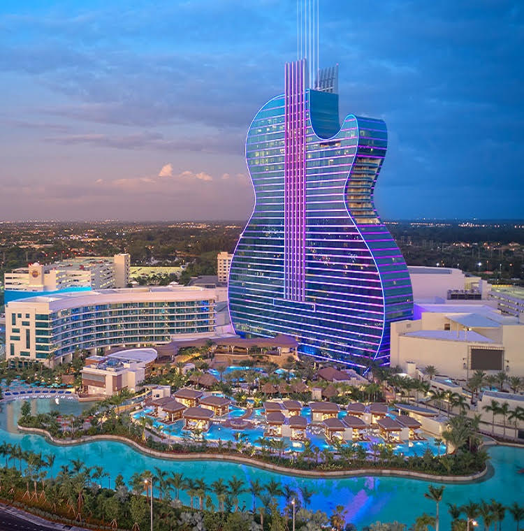 A large hotel building shaped like a guitar is prominently featured, with vibrant lights illuminating its exterior. The surrounding area includes a lagoon-style pool with palm trees and cabanas, creating a relaxing atmosphere. Soft colors are present in the sky as daylight transitions to evening.