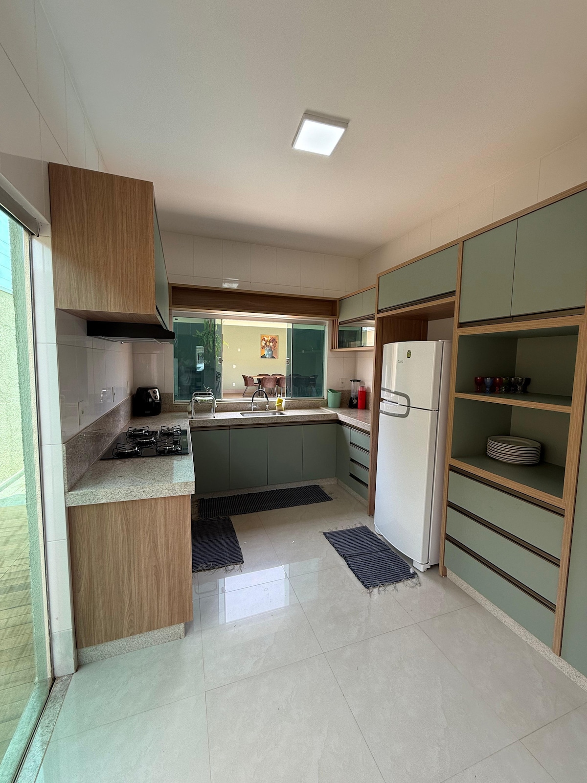 The kitchen features modern cabinetry in soft green tones, complemented by a light countertop. Stainless steel appliances, including a refrigerator and a stovetop, are visible. Natural light enters through a large window, illuminating the functional cooking space with accessories neatly arranged on open shelves.