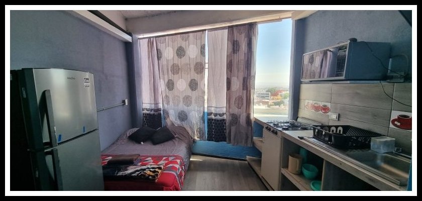 A studio apartment features large windows with sheer curtains allowing natural light to fill the space. A spacious bed is positioned next to a kitchenette equipped with essential appliances, including a refrigerator and a stove. The overall layout promotes a functional and comfortable living area.