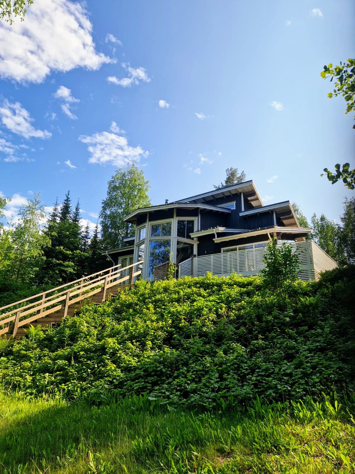 The modern cabin is situated on a hill, surrounded by lush greenery. A wooden staircase leads to the entrance, while large windows reflect natural light, showcasing the structure's inviting design. The sky is bright, dotted with clouds, enhancing the serene atmosphere.