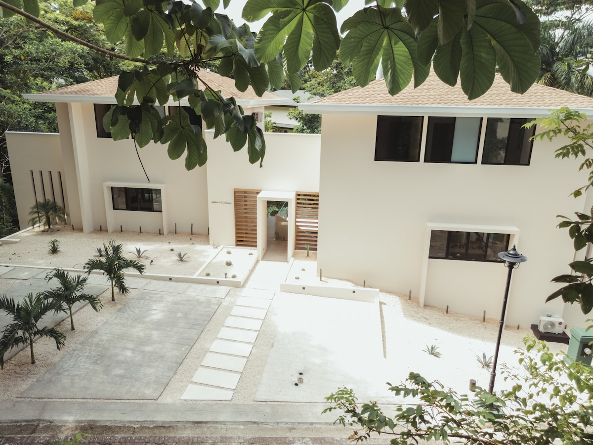 The exterior of a contemporary building is presented, showcasing a clean white façade with strategically placed windows. A landscaped pathway leads to the entrance, bordered by minimalistic greenery. The surrounding area is well-maintained, inviting a sense of tranquility amidst the lush environment.