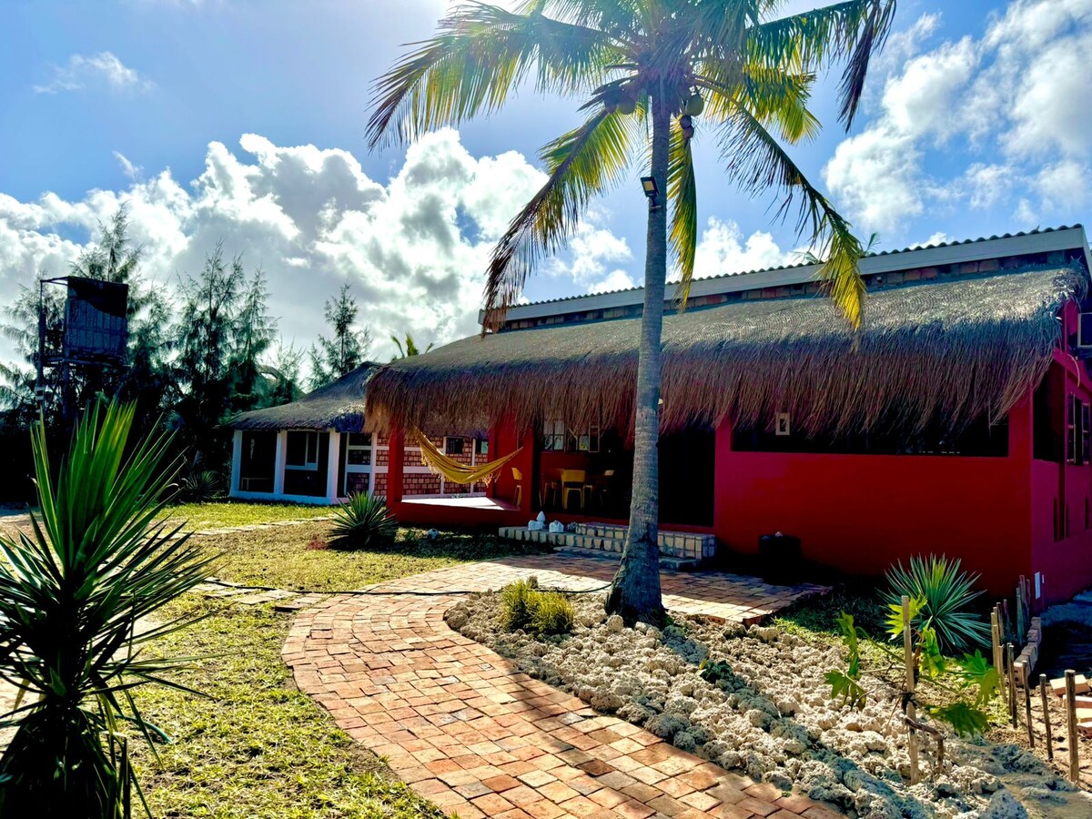 A vibrant red house with a thatched roof is set within a lush garden. A winding brick pathway leads to its entrance. Palm trees and low shrubs surround the area, creating a serene outdoor environment that invites relaxation.