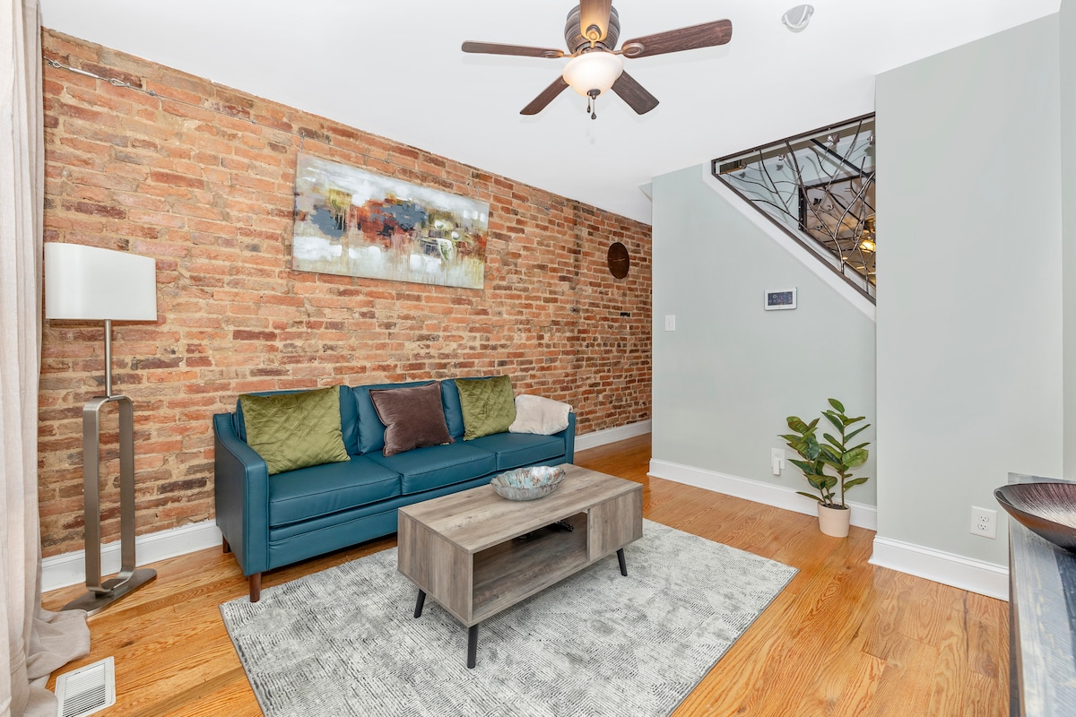 A cozy living area is highlighted by exposed brick walls and hardwood flooring. A teal sofa is adorned with pillows, positioned across from a wooden coffee table on a soft area rug. Natural light filters through a nearby window, illuminating the space.