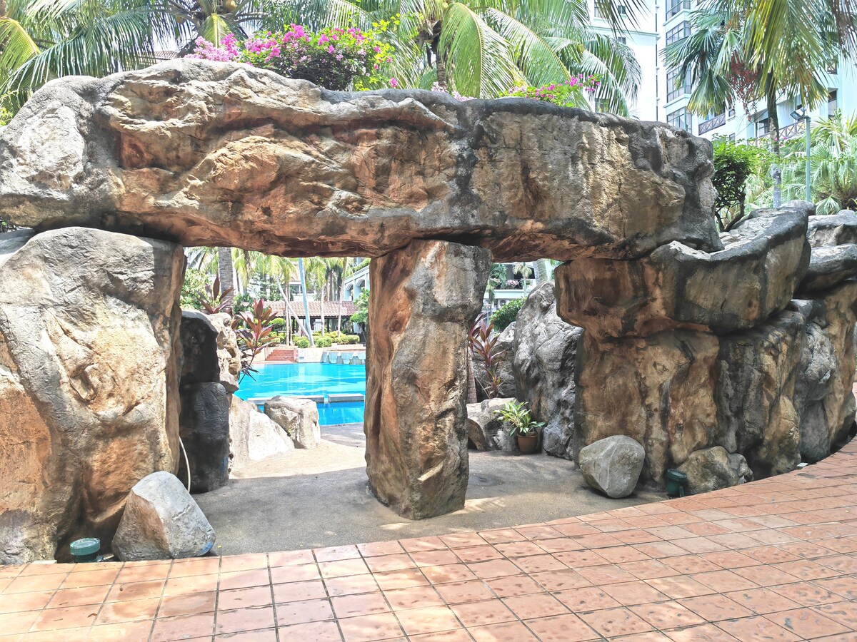 A natural stone archway is framed by tropical plants, leading toward a swimming pool visible in the background. The textured rocks add a rustic charm, while colorful flowers provide a lively touch to the serene outdoor space.