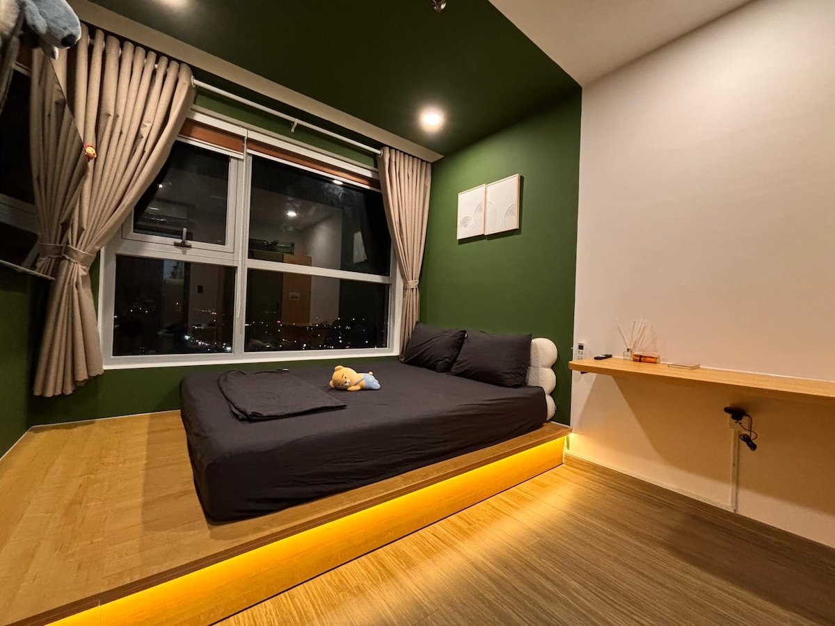 A cozy sleeping area features a spacious bed with a dark cover placed on a raised platform. Natural light enters through the large window adorned with soft curtains. A minimalist desk is positioned against the wall, adding to the room's functionality.