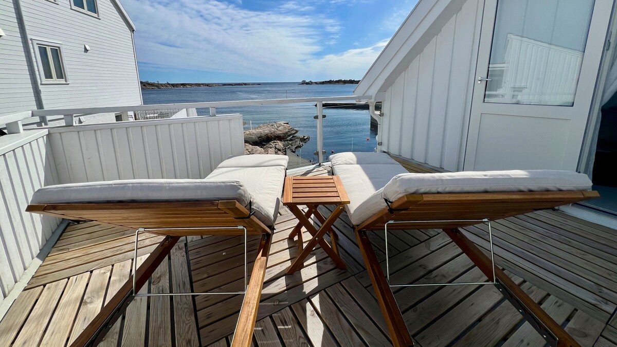 Two chaise lounge chairs are positioned on a sunlit deck, facing a serene view of the sea. A small table rests nearby, complementing the seating area. The peaceful atmosphere is enhanced by the surrounding wooden structure and clear blue skies.
