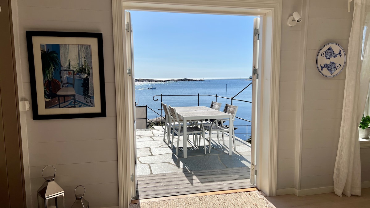 A view from the open doorway reveals a bright patio furnished with a dining table and chairs, overlooking the calm sea. The sunlight reflects off the water, enhancing the serene setting. A framed artwork is visible on the wall beside the door.