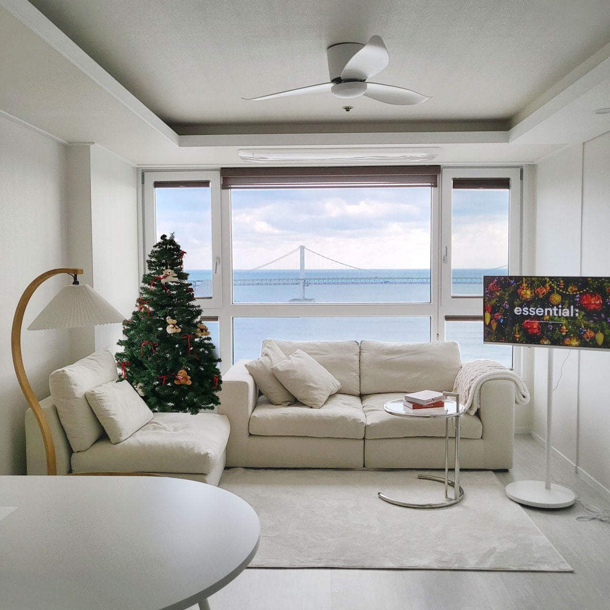 A modern living area features a white sofa arranged beside a table and a decorative Christmas tree. Large windows provide a view of the ocean and a bridge. A television stands on a stand beside the sofa, enhancing the room's light and airy feel.