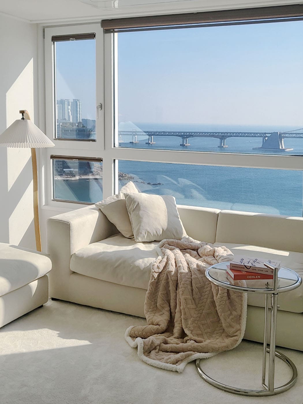 A modern living area features a white sofa adorned with plush cushions and a soft throw. A circular glass table displays neatly stacked books. Large windows offer expansive views of the ocean and a distant bridge, allowing natural light to fill the space.