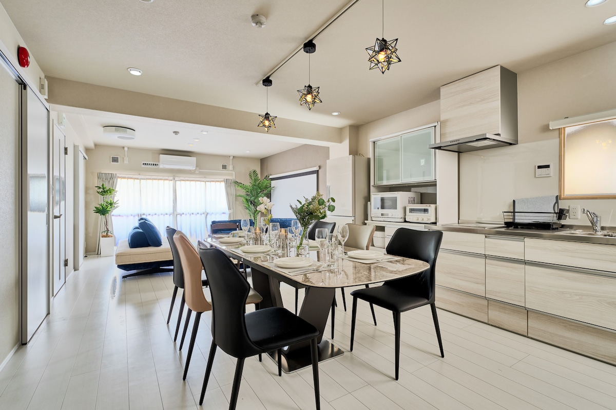 A modern dining area includes a large table set for eight. Sleek black and beige chairs surround the table, while stylish pendant lights hang above. A well-equipped kitchen features contemporary cabinetry and appliances, with natural light filtering through large windows adorned with sheer drapes.