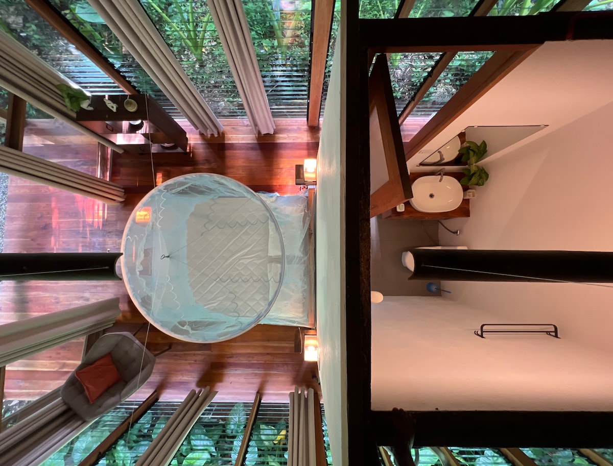 An aerial view of the bedroom reveals a spacious bed draped with a protective canopy, surrounded by large glass windows that invite natural light and offer views of the surrounding greenery. A modern bathroom area is visible, featuring clean lines and supporting elements like a sink and shelving.