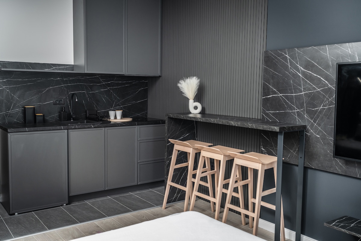 An elegant kitchen area is highlighted with sleek dark cabinetry and a black marble backsplash. A minimalist wooden bar with three stools adds functionality, while decorative accents are positioned on the countertop. The space exhibits a modern and sophisticated design.
