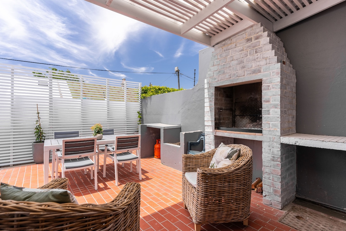 An outdoor deck area features a built-in braai with a brick surround. A dining table is paired with four chairs, while two wicker chairs provide seating. Potted plants add a touch of greenery, and the space is illuminated by natural light under a slatted roof.