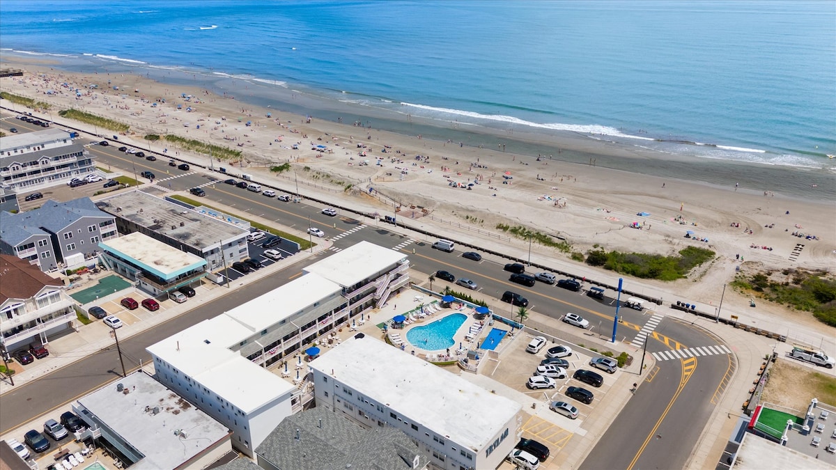 An aerial view captures the coastline with a sandy beach and numerous sunbathers. A community pool is visible near the center of the complex, surrounded by parked vehicles. The ocean's blue waters stretch out toward the horizon, presenting a serene coastal setting.