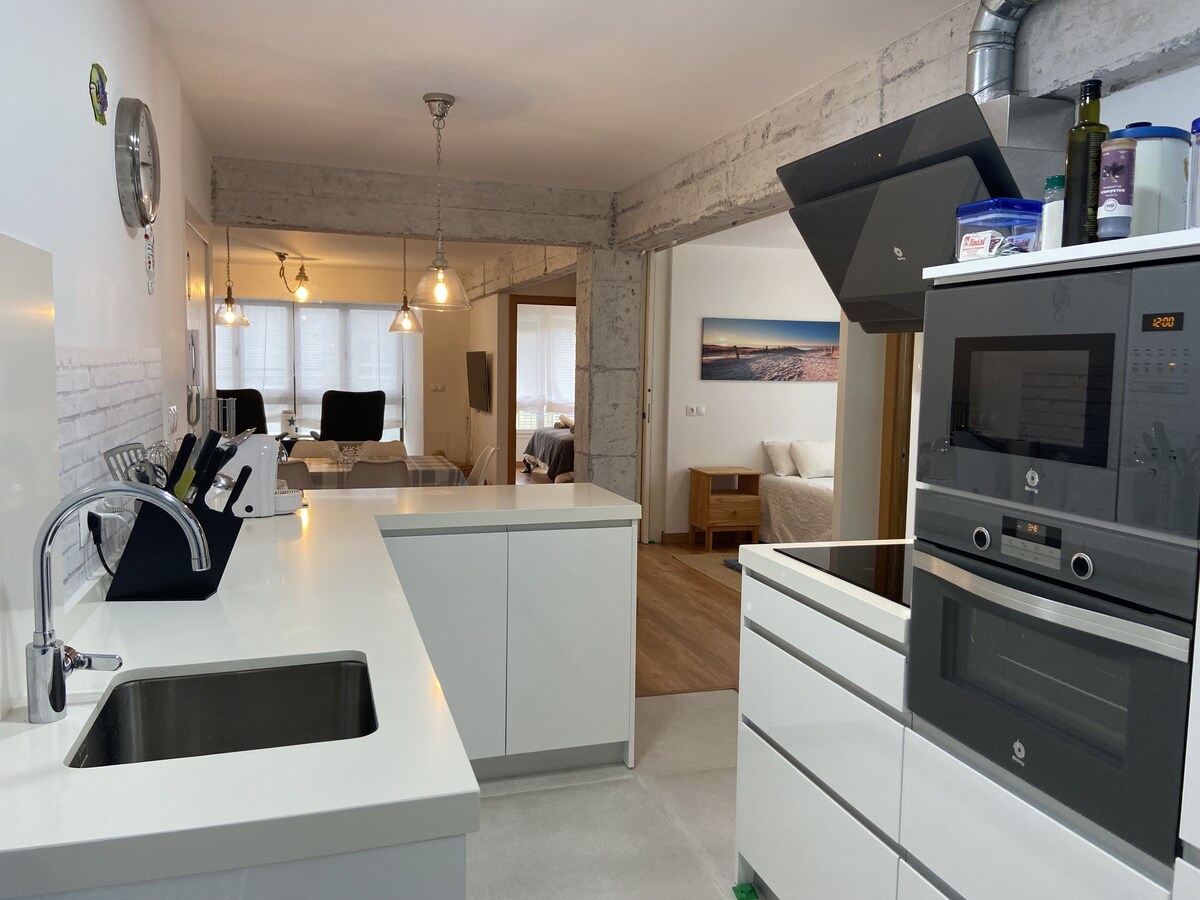 A modern kitchen is presented with sleek white cabinetry and a spacious countertop. An integrated sink and oven are visible, along with minimalist lighting fixtures. The open layout connects to a living area, allowing for a bright and airy atmosphere.