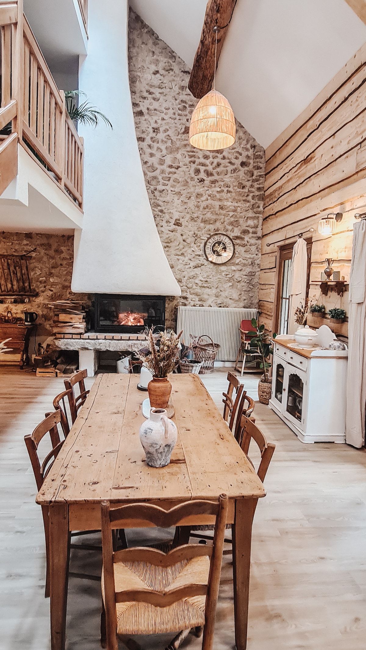 A large dining table crafted from natural wood is positioned centrally in an airy space. The high ceiling features wooden beams, and a modern fireplace is set against a stone wall. Natural light filters through nearby windows, enhancing the inviting setting.