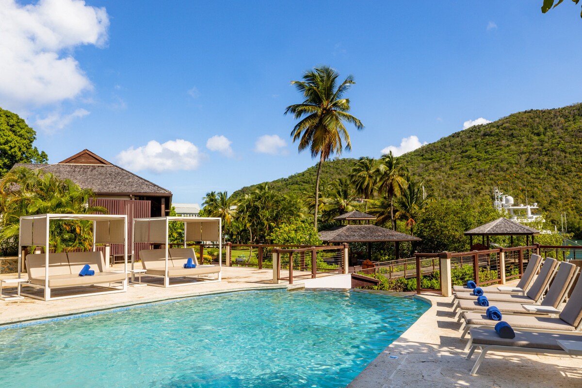 A clear blue swimming pool is surrounded by lounge chairs and shaded cabanas, with vibrant palm trees framing the scene. Lush greenery and distant hills provide a serene backdrop, highlighting a tranquil setting for relaxation.