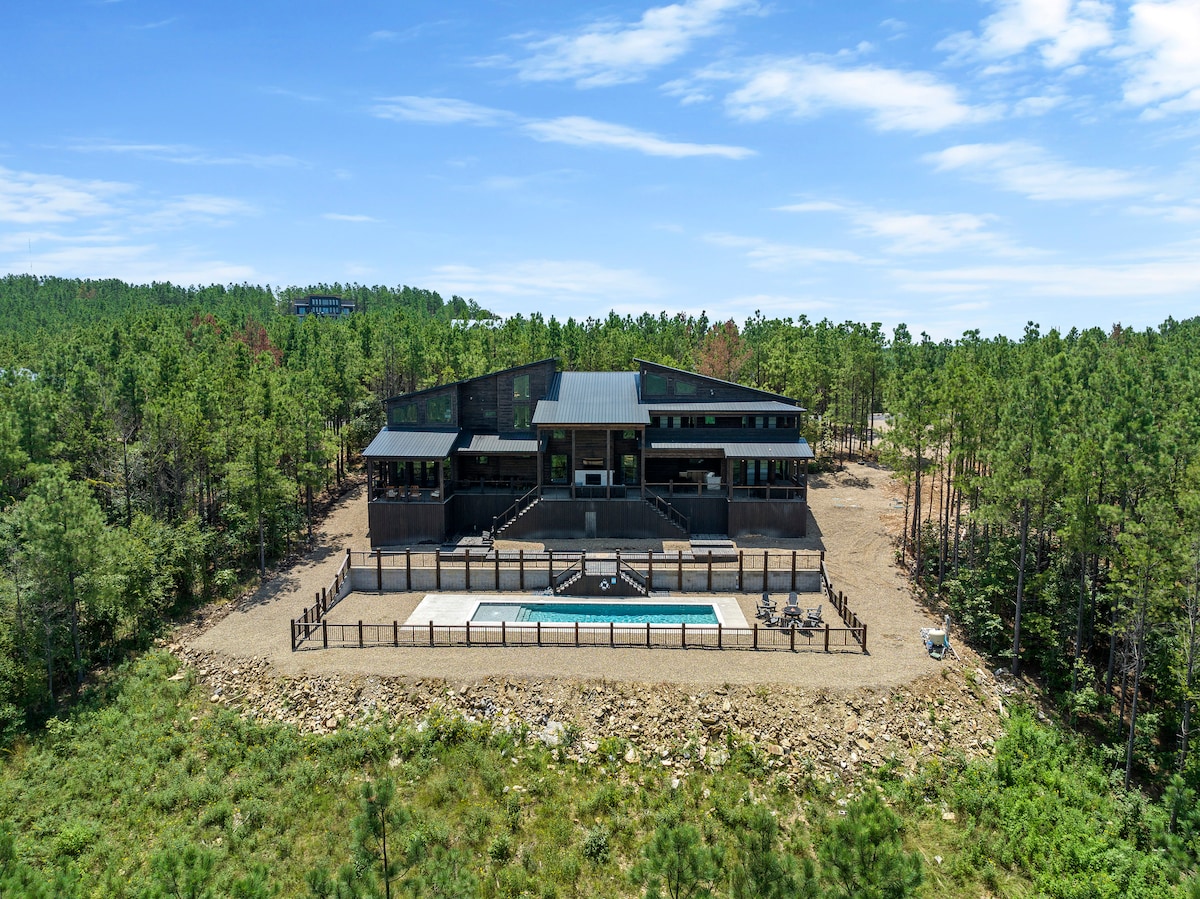 A modern two-story home is situated among lush greenery, featuring a spacious outdoor area. A fenced heated pool is visible in the foreground, surrounded by natural landscaping. The expansive windows of the house reflect the surrounding trees, enhancing the connection with nature.