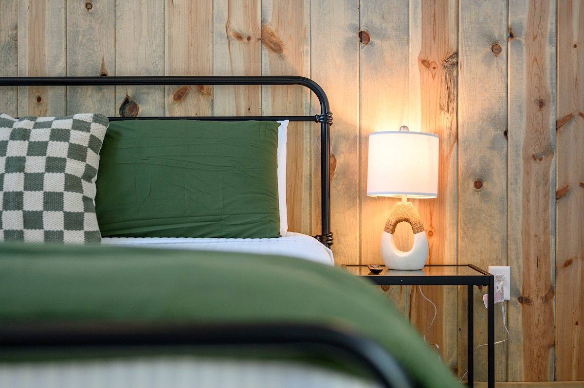 Primer plano de mesita de noche con lámpara junto a una cama de temática verde contra una pared con paneles de madera.