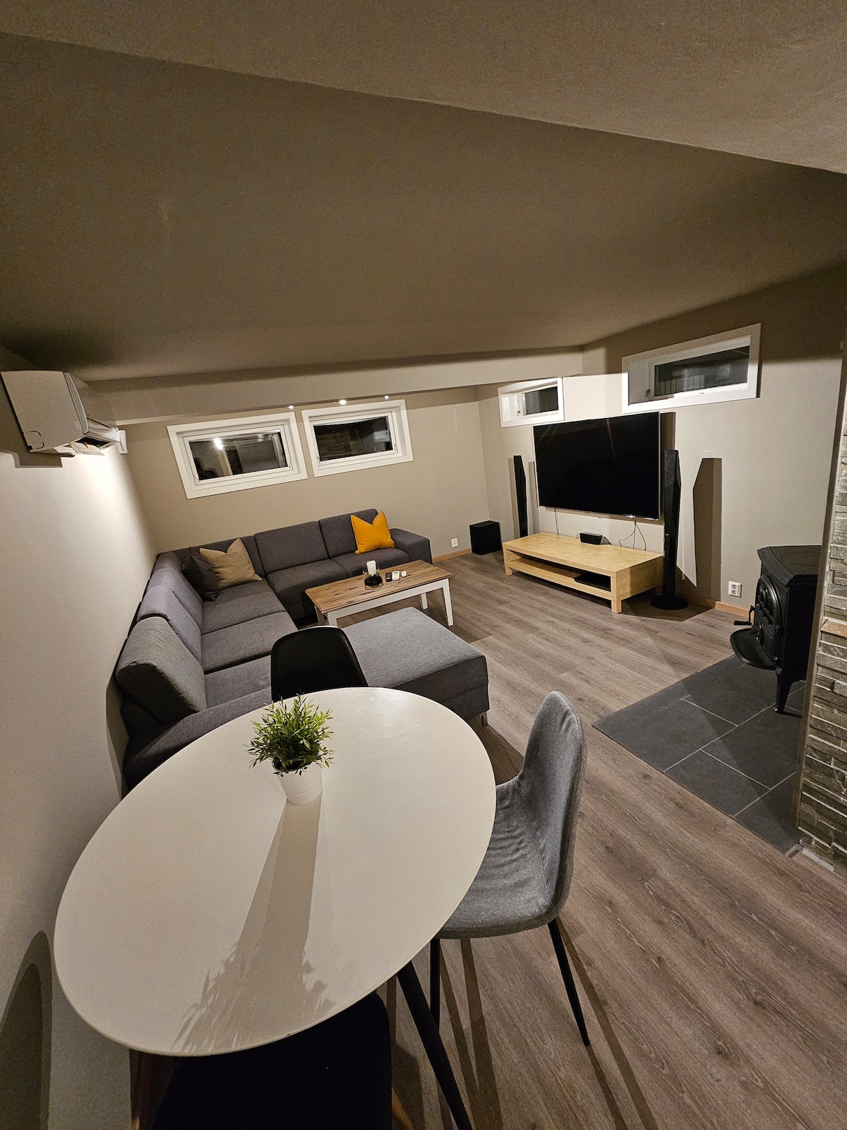 The living area is designed with a sectional sofa, arranged around a flat-screen television. A table with four chairs occupies one corner, while large windows provide natural light. The neutral walls and modern flooring enhance the space's contemporary feel.
