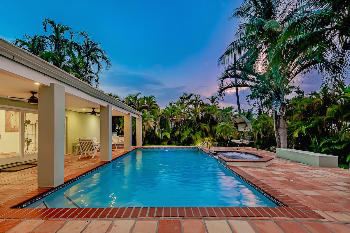 A serene pool area is surrounded by lush tropical landscaping, creating a tranquil atmosphere. The clear water of the pool reflects the evening sky, while a jacuzzi is positioned nearby. A covered patio provides shaded seating for relaxation and enjoyment.