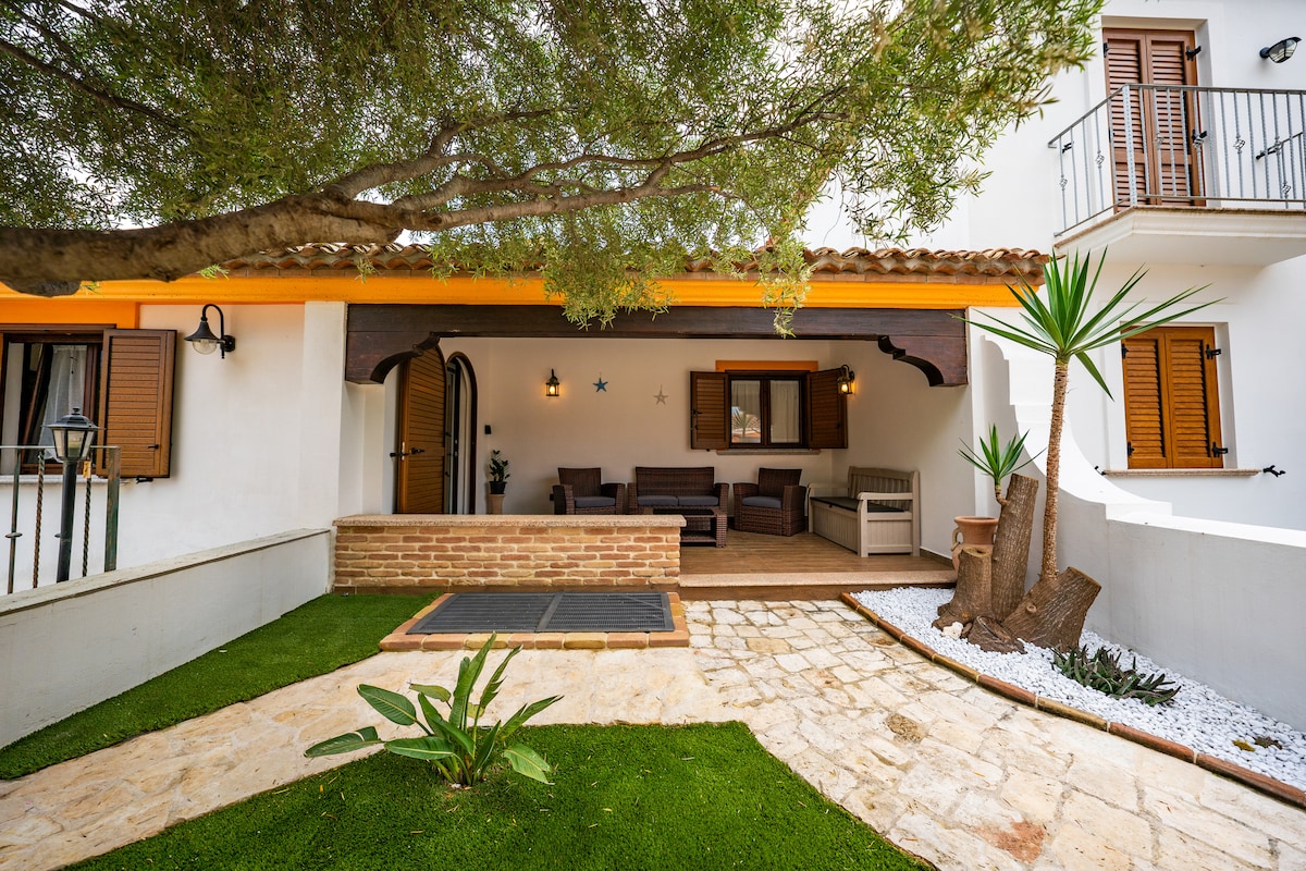 An outdoor area features a welcoming covered veranda with comfortable seating, surrounded by a lush garden with synthetic grass. A decorative stone path leads through the space, complemented by potted plants and white gravel, enhancing the tranquil ambiance.