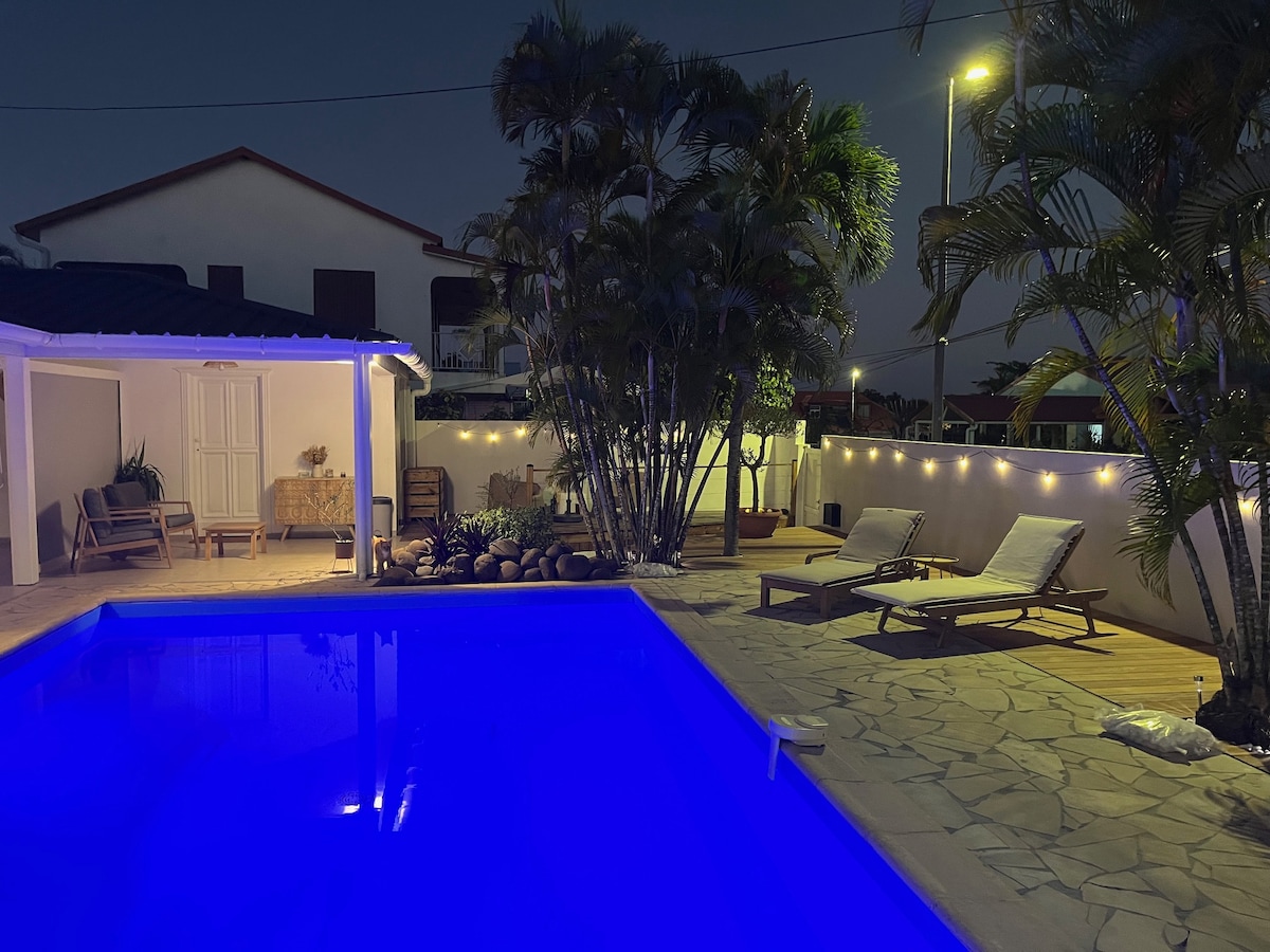 A tranquil private pool area is illuminated at night, featuring a bright blue pool surrounded by stone decking. Palm trees create a lush backdrop, with comfortable lounge chairs positioned nearby. Soft lighting adds ambiance to the relaxation space, enhancing nighttime enjoyment.