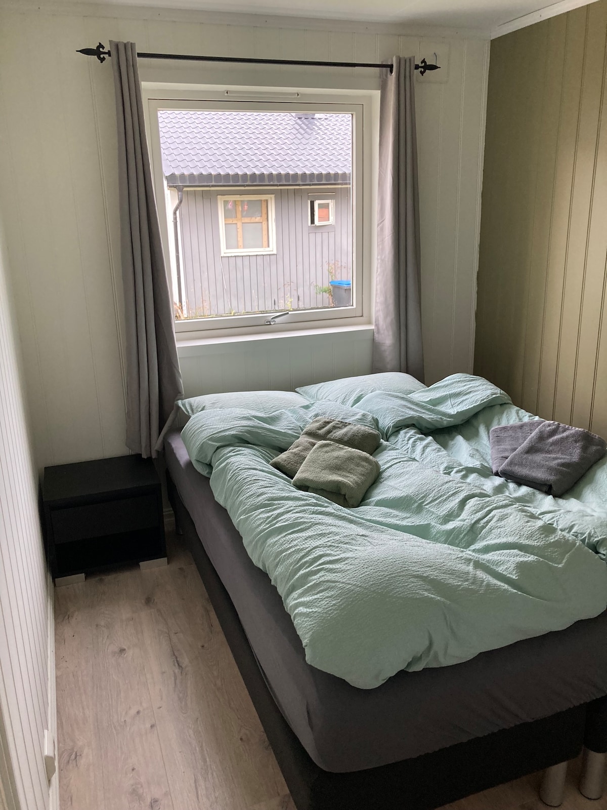 A compact sleeping area features a spacious bed with soft bedding in light green hues. Folded towels are neatly arranged on the bed. A window allows natural light to fill the room, framed by light grey curtains. A small bedside table is positioned beside the bed.