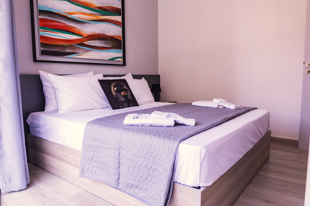 A spacious bedroom is featured, showcasing a large bed with crisp white linens and a textured gray blanket. A striking piece of wall art adds visual interest. Soft natural light fills the room, accentuating the warm tones of the wooden floor and modern furnishings.