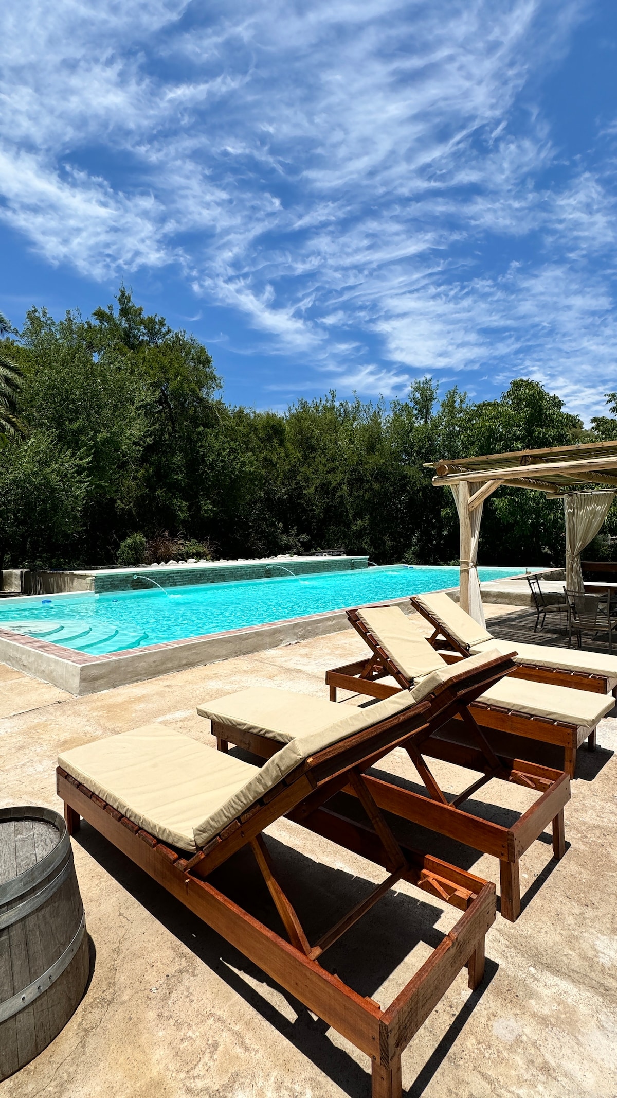 A tranquil outdoor area features wooden loungers arranged beside a refreshing swimming pool. Lush greenery surrounds the pool, and a shaded gazebo offers a space for relaxation. The clear blue sky adds to the serene atmosphere, inviting guests to unwind.