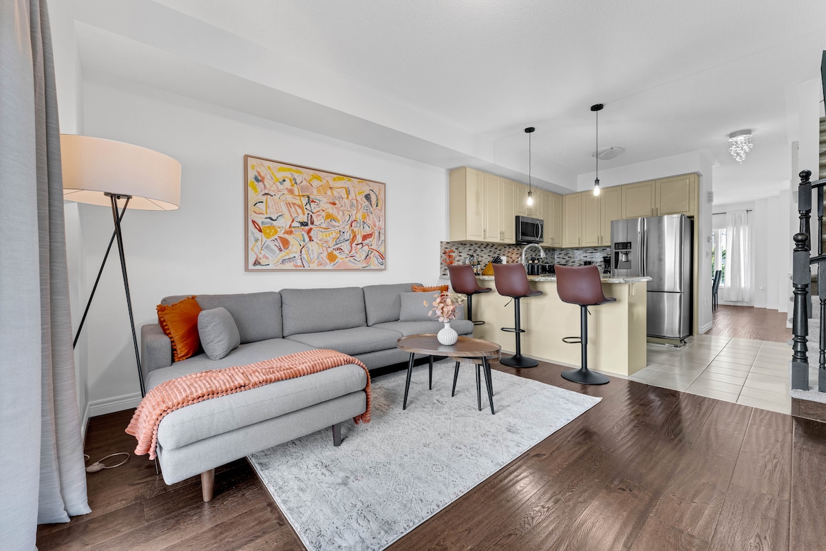 The living area features a light grey sectional sofa with an orange throw and patterned cushion, paired with a wooden coffee table. An art piece adorns the wall, while modern bar stools surround the kitchen island in the background, enhancing the open layout.